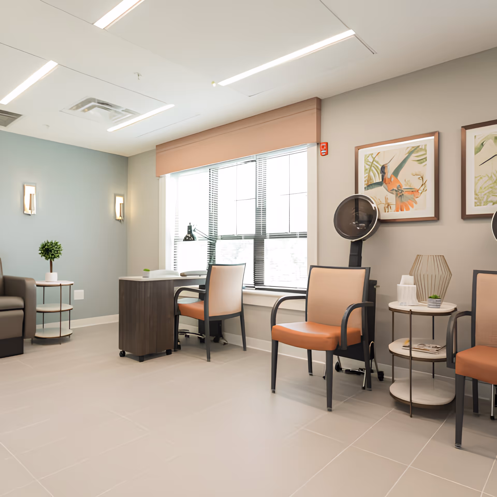 Bright senior living common room with seating, small tables, a desk by a window, and framed wall art.