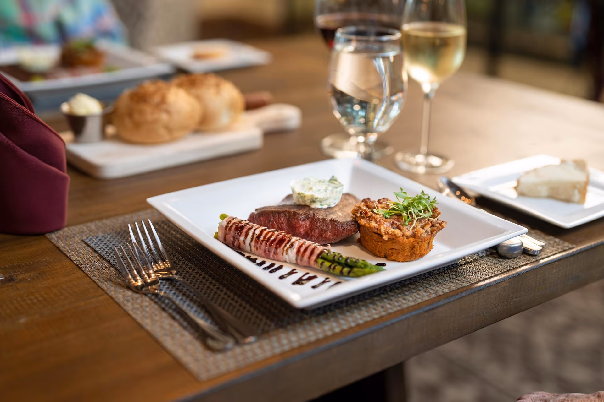 A plated gourmet meal on a wooden table featuring a piece of steak topped with herb butter, a bacon-wrapped asparagus spear, and a savory muffin garnished with microgreens. In the background, there are two bread rolls on a cutting board, a slice of bread on a small plate, and glasses of water and white wine.