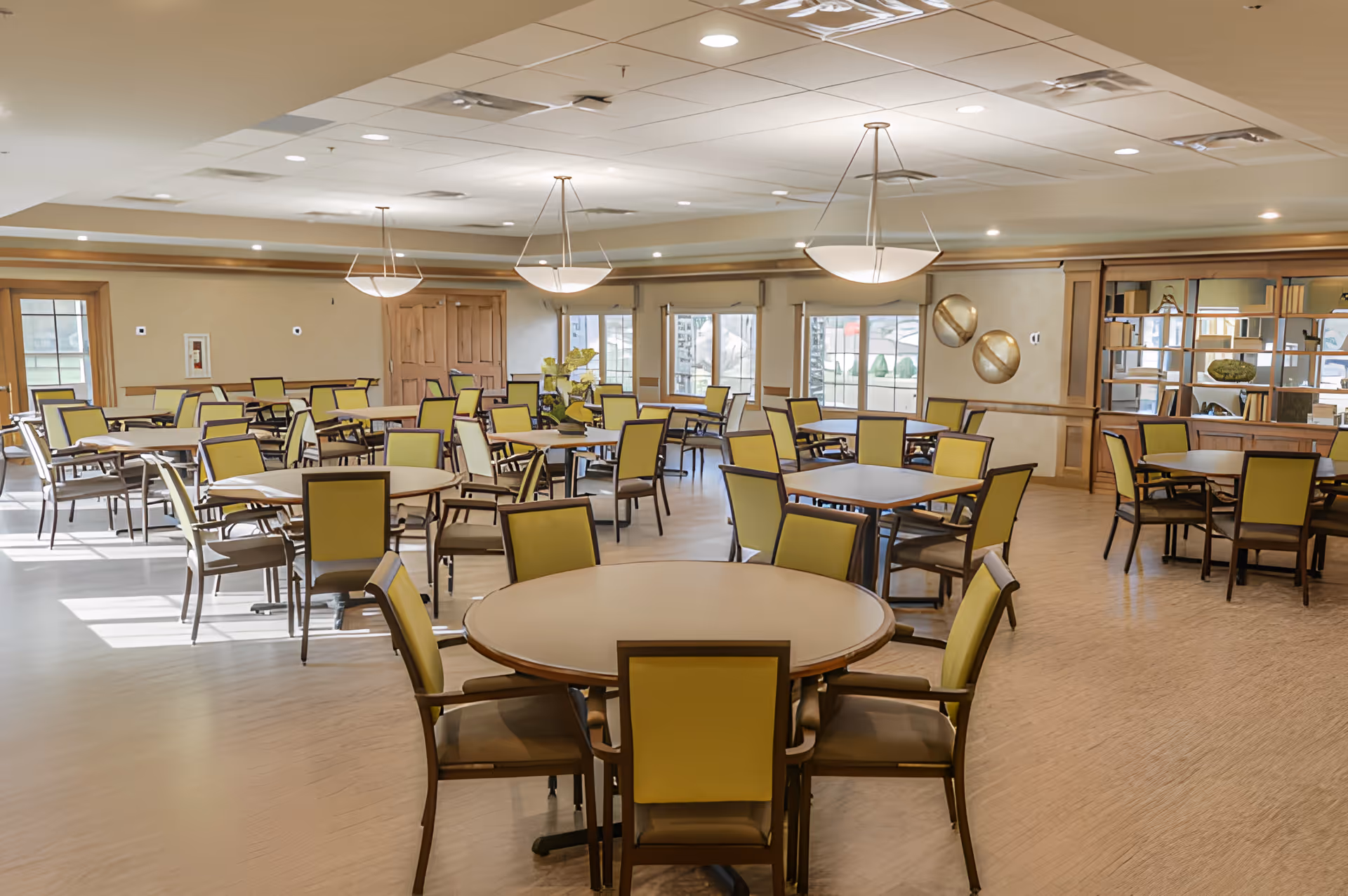 A spacious dining room with multiple round and square tables surrounded by chairs with yellow backs and brown frames. The room has large windows letting in natural light, modern ceiling lights, and light-colored flooring. There are decorative elements on the walls and a mirrored shelving unit on one side.