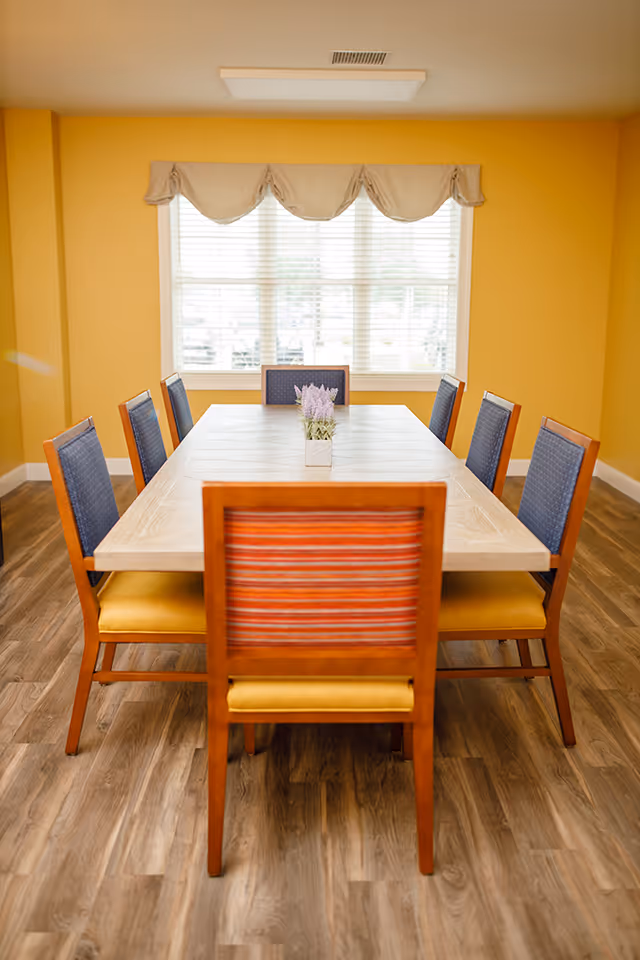 A dining room with a rectangular wooden table surrounded by eight chairs. The chairs have wooden frames with yellow seats and blue or striped backs. A small potted plant with purple flowers is centered on the table. The room has yellow walls, a large window with white blinds and a beige valance, and wood-patterned flooring.