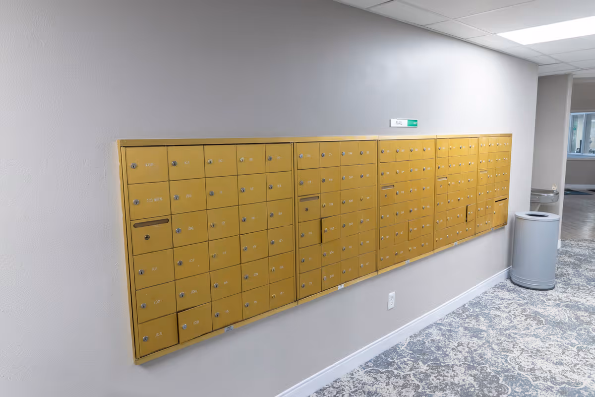 A wall-mounted set of yellow mailboxes with multiple locked compartments in a hallway with patterned carpet and a trash can nearby.