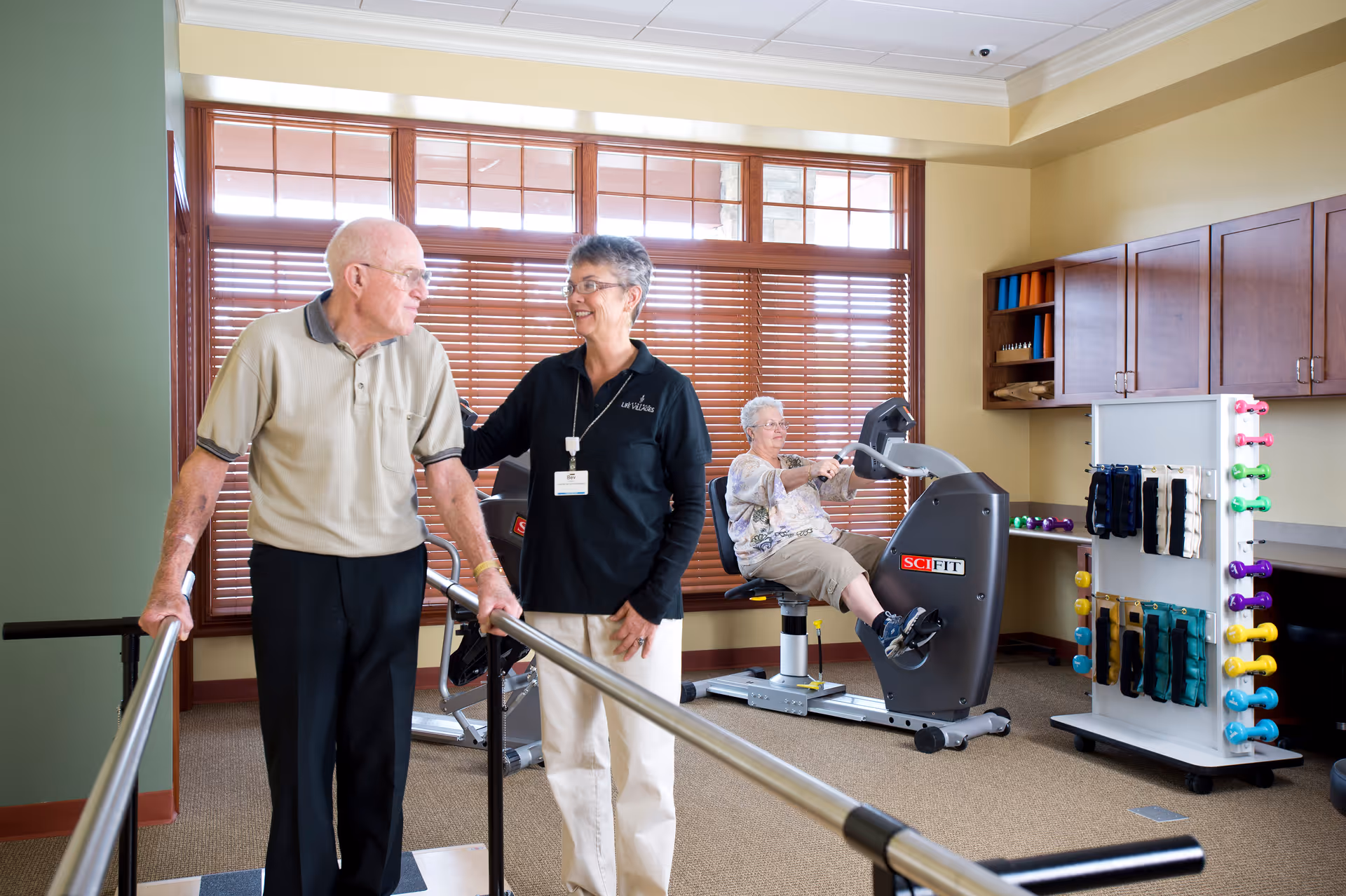 An elderly man uses parallel bars for walking exercise with the assistance of a female staff member in a fitness room. In the background, an elderly woman is seated on a recumbent exercise bike. The room has large windows with wooden blinds, exercise equipment, and a rack with colorful dumbbells and ankle weights.