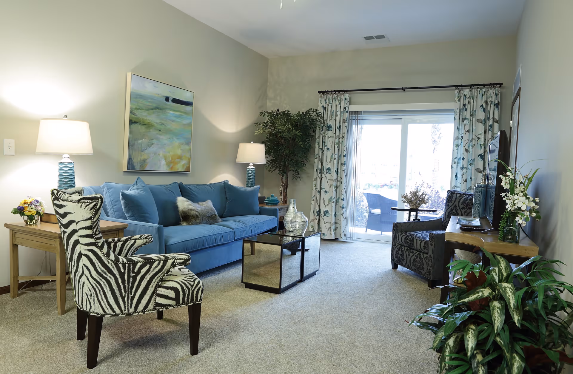 A bright living room with a blue couch, a zebra-patterned armchair, a patterned armchair, two wooden side tables with lamps, a glass coffee table, a TV on a wooden stand, floral curtains, and a sliding glass door leading to an outdoor patio. There are also several plants and a painting on the wall.