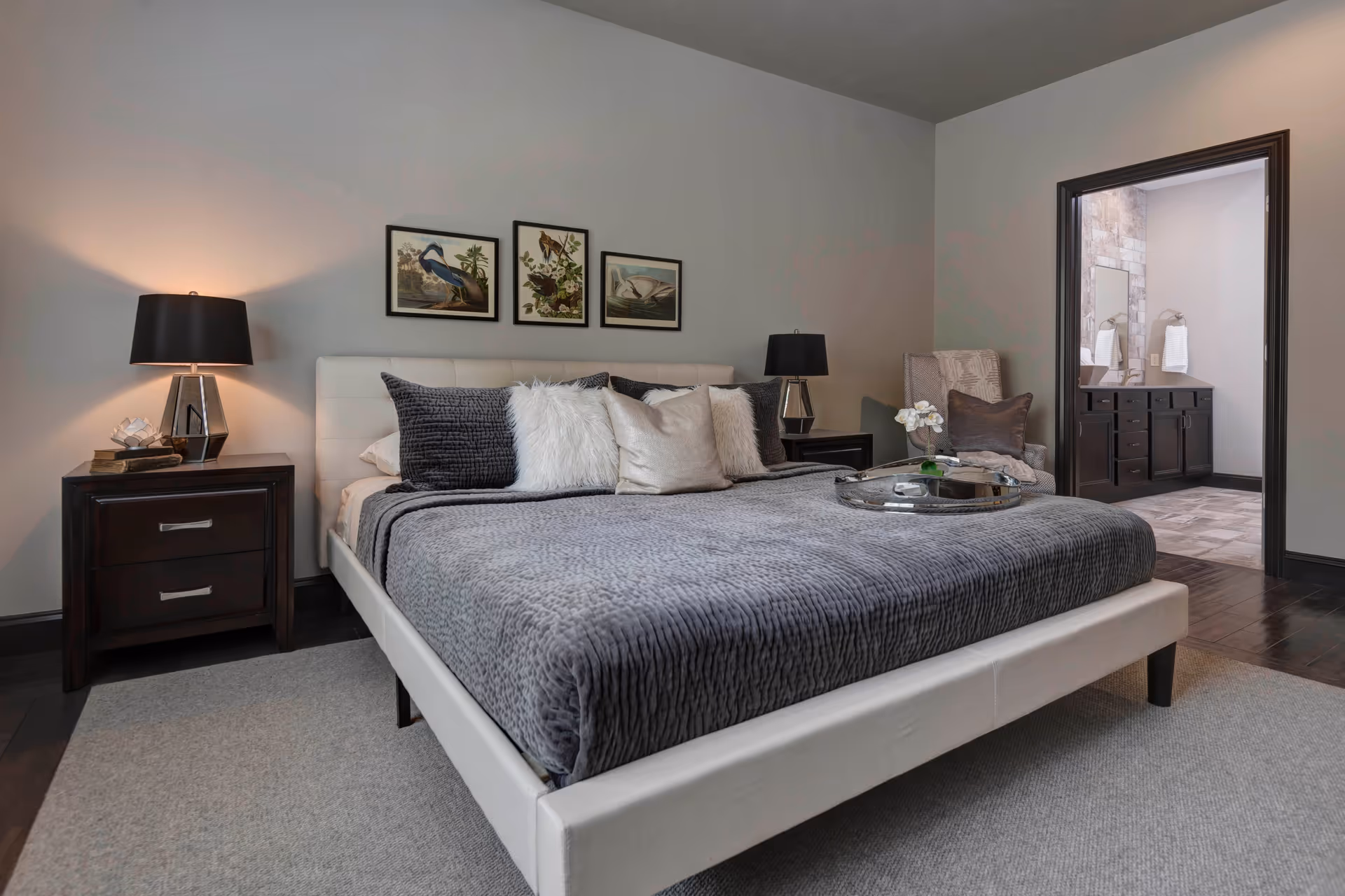 Bedroom with a gray upholstered bed and bedding, two nightstands with lamps, wall art above the headboard, and an open doorway to a bathroom.
