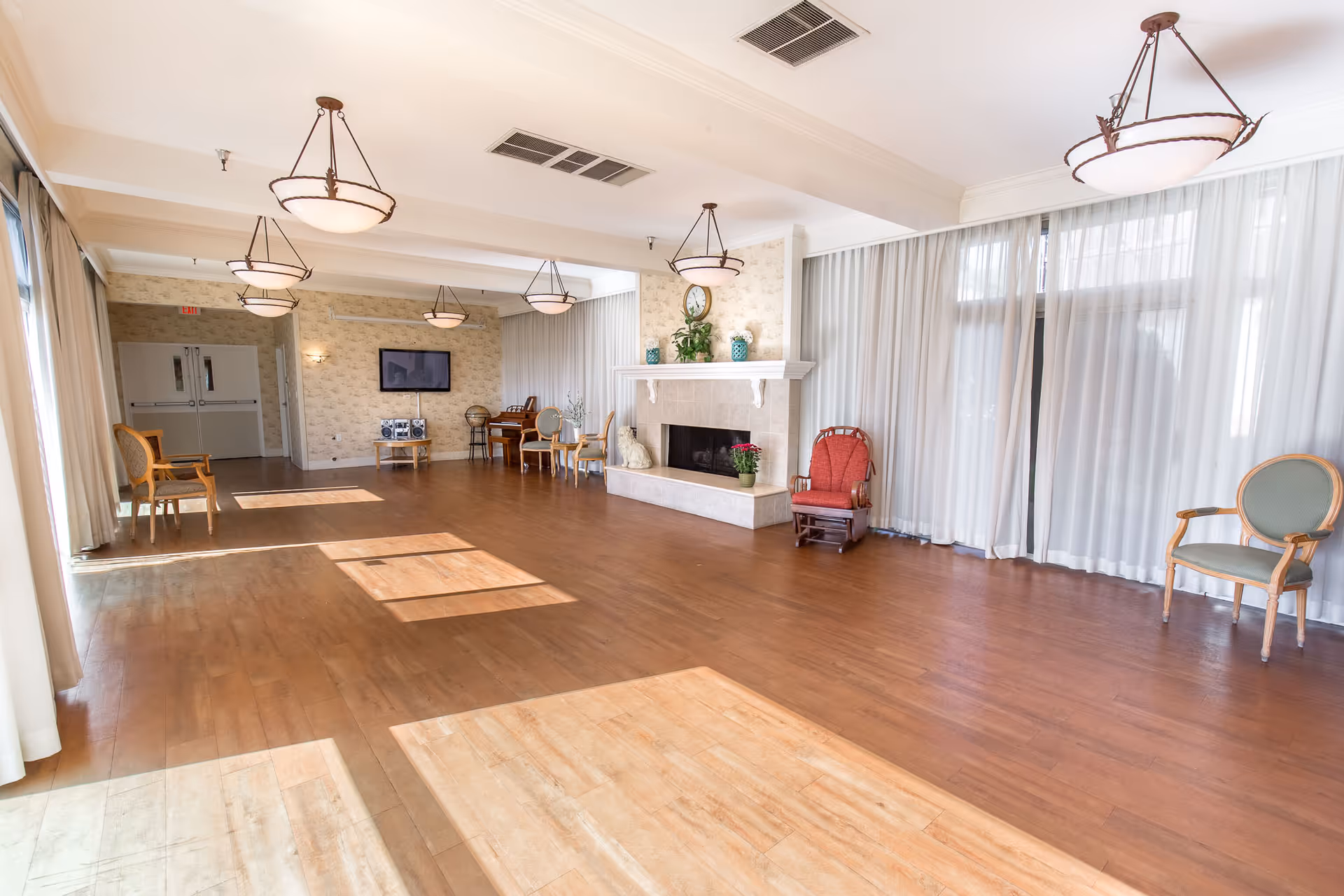 A spacious, well-lit common area with wooden flooring, several chairs arranged along the walls, a fireplace with decorative items on the mantel, large windows with sheer curtains, and ceiling light fixtures.