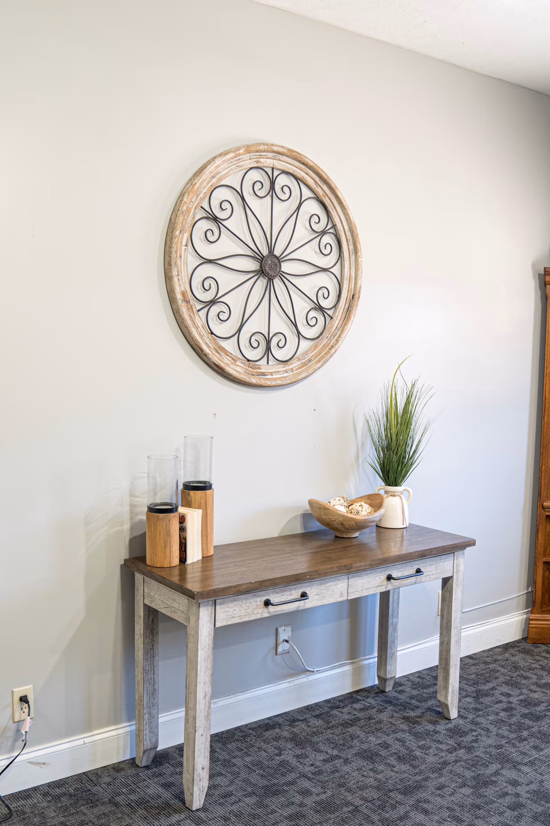 A wooden console table with two drawers against a light gray wall. On the table are two tall glass candle holders with wooden bases, a wooden bowl with decorative balls, and a white vase with green grass-like plants. Above the table hangs a round decorative wall piece with a wooden frame and black metal scrollwork design. The floor is carpeted in a dark pattern.