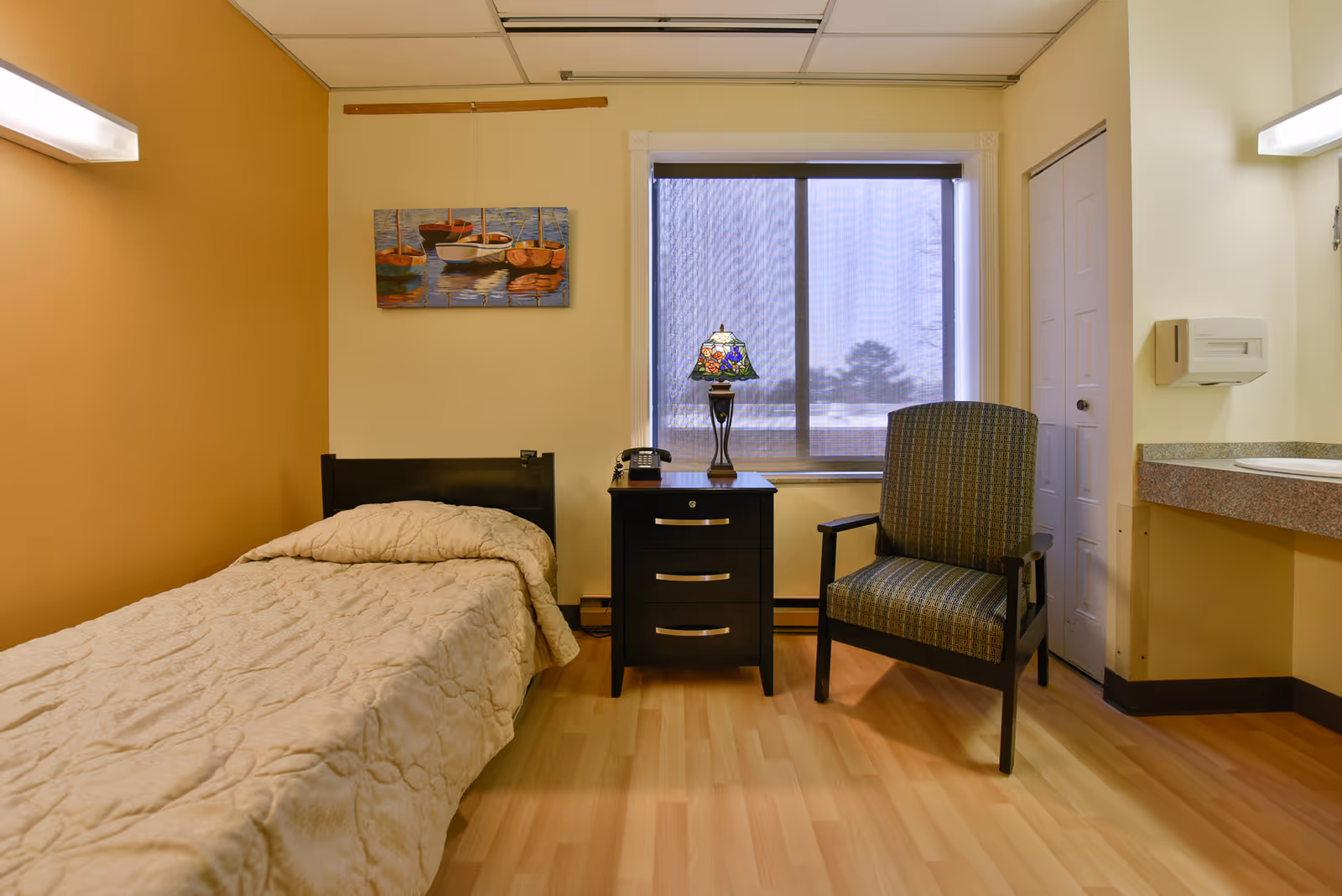 A small, cozy bedroom in a nursing facility with a single bed covered in a beige quilt, a black nightstand with three drawers, a decorative lamp, and a telephone. Next to the nightstand is a cushioned armchair with a patterned fabric. A window with a mesh screen is behind the nightstand and chair. The walls are painted in warm tones, and there is a painting of boats hanging above the bed. A sink with a paper towel dispenser is visible on the right side of the room.