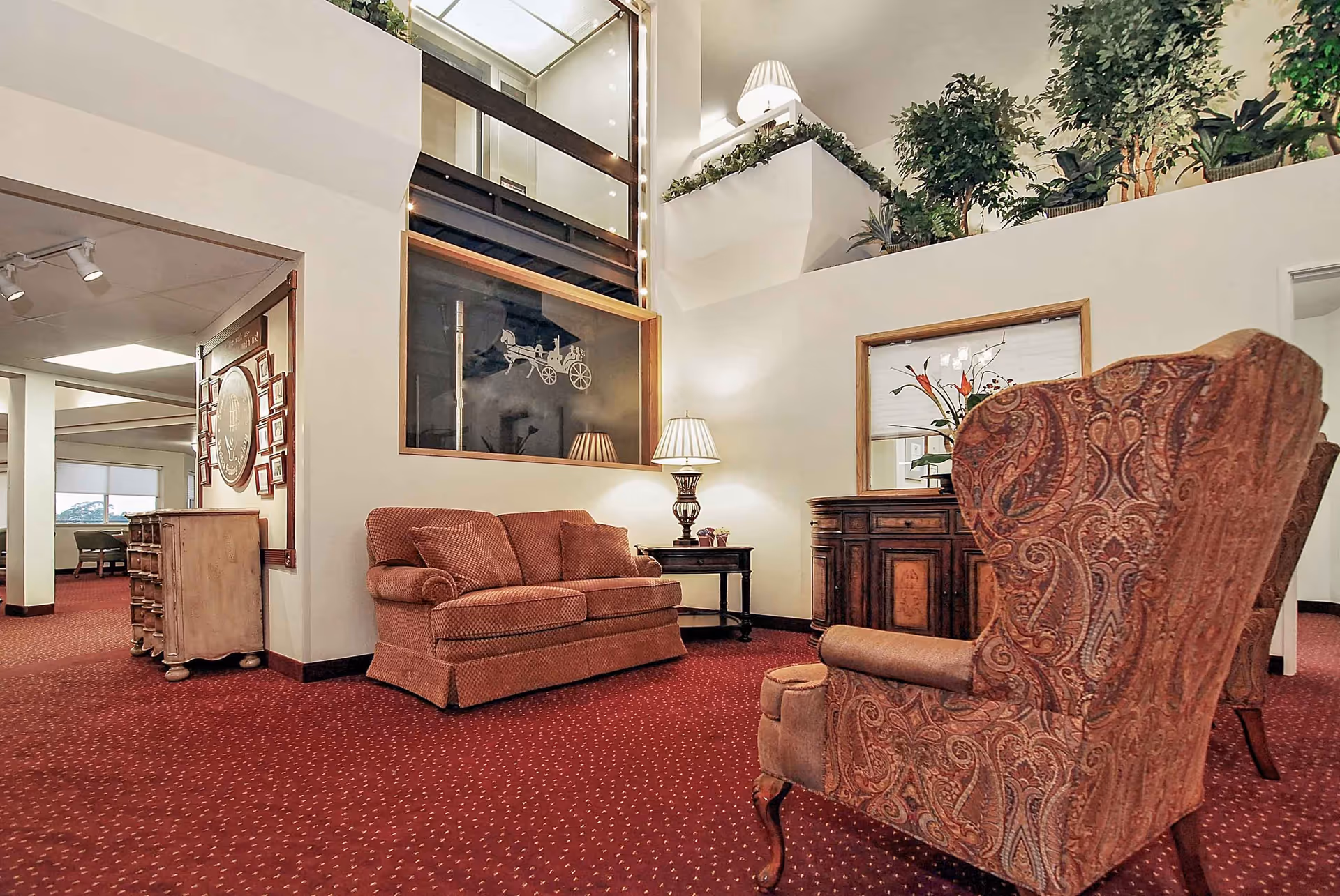A cozy sitting area in a senior living facility with a patterned armchair, a cushioned loveseat, a wooden side table with a lamp, and a wooden cabinet with a mirror above it. The room has red carpet flooring, white walls, and several green plants on a ledge above. There is a large window with a decorative horse and carriage design and a hallway leading to another room in the background.