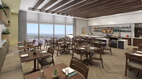 A spacious dining room with multiple wooden tables and chairs arranged neatly. The room features large windows with a scenic view of the city and mountains in the distance. There is a long cushioned bench along one wall, shelves with plants, and a counter area with a coffee machine and other amenities. The ceiling has exposed wooden beams and the floor has a herringbone pattern.