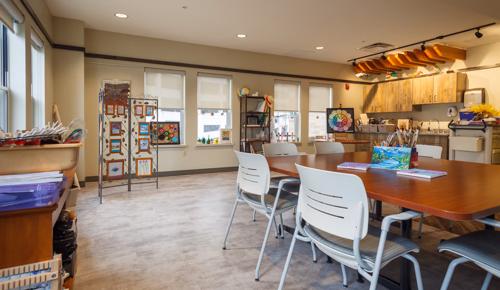 A bright and spacious activity room with a large wooden table surrounded by white chairs. The room has several windows with blinds, a display rack with artwork, shelves with books and decorations, and a kitchenette area with wooden cabinets and art supplies. The ceiling features a wooden beam with colorful bird decorations.