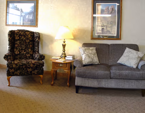 Seating area with a patterned armchair, small wooden side table with a lamp and books, and a two-seat sofa beneath framed pictures.