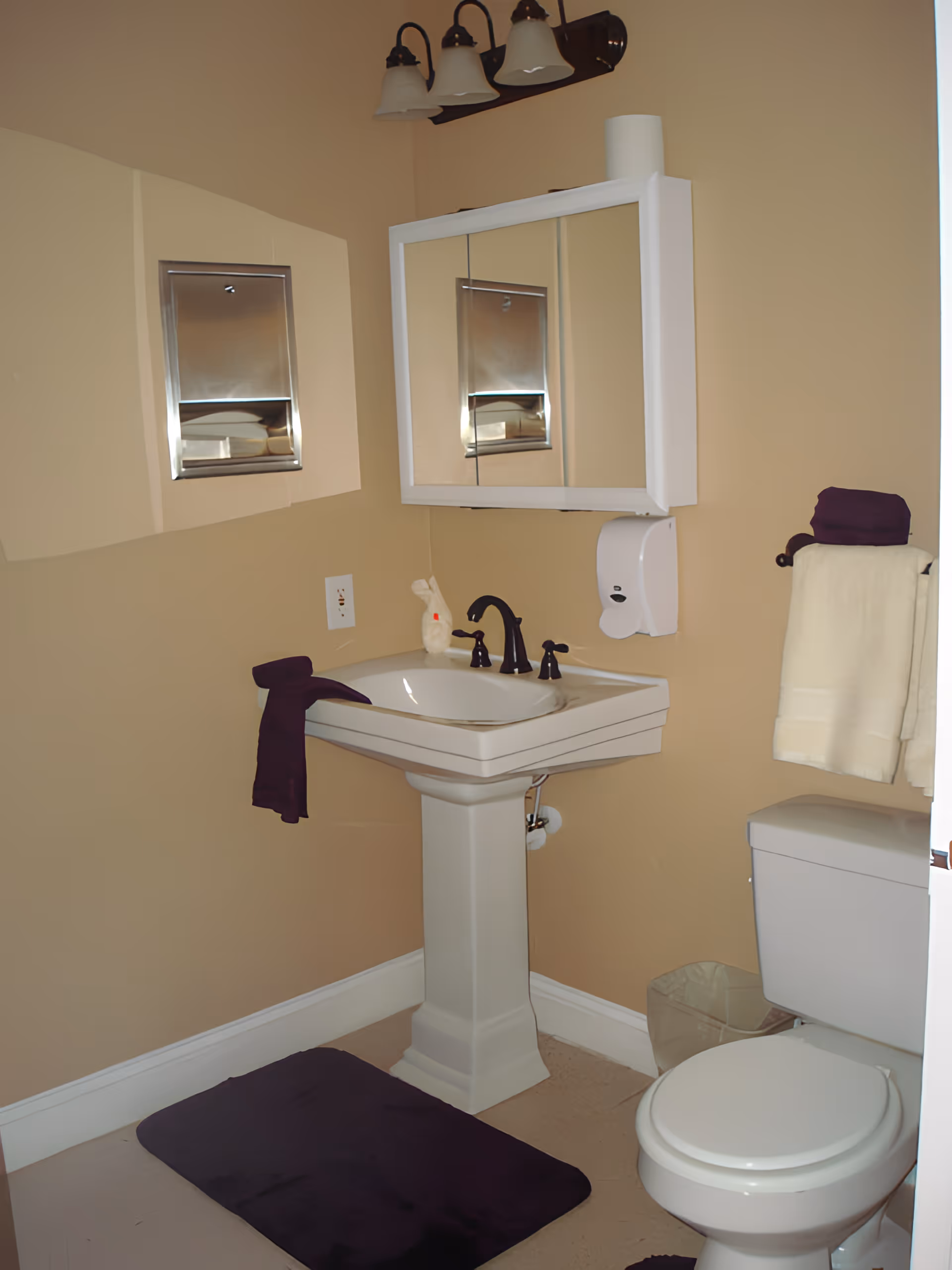 A small bathroom with beige walls featuring a white pedestal sink with dark bronze faucet, a mirrored medicine cabinet above the sink, a toilet to the right, a towel rack with white and dark purple towels, a dark purple bath mat on the floor, and a wall-mounted soap dispenser.