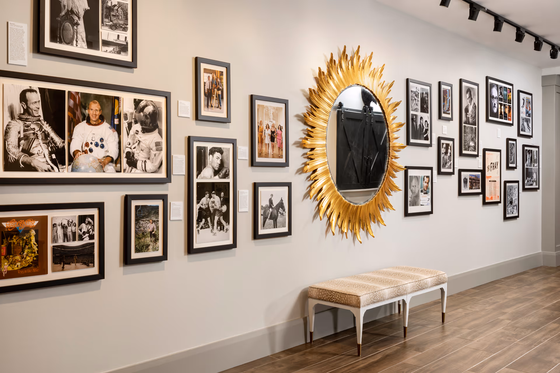 Hallway gallery with multiple framed photographs, a gold sunburst mirror, and a bench against a neutral wall.