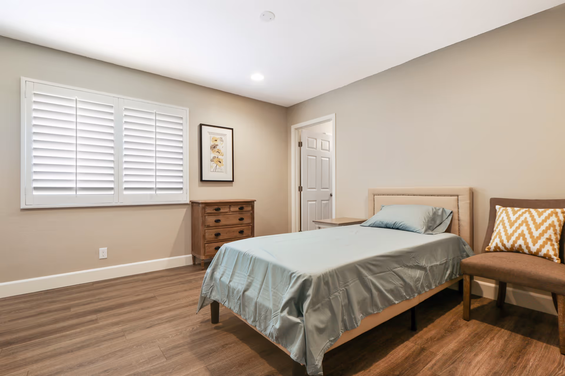 A simple, clean bedroom with a single bed covered in light blue bedding, a wooden dresser against the wall, a chair with a yellow and white chevron-patterned pillow, a window with white shutters, and a framed floral picture on the wall.