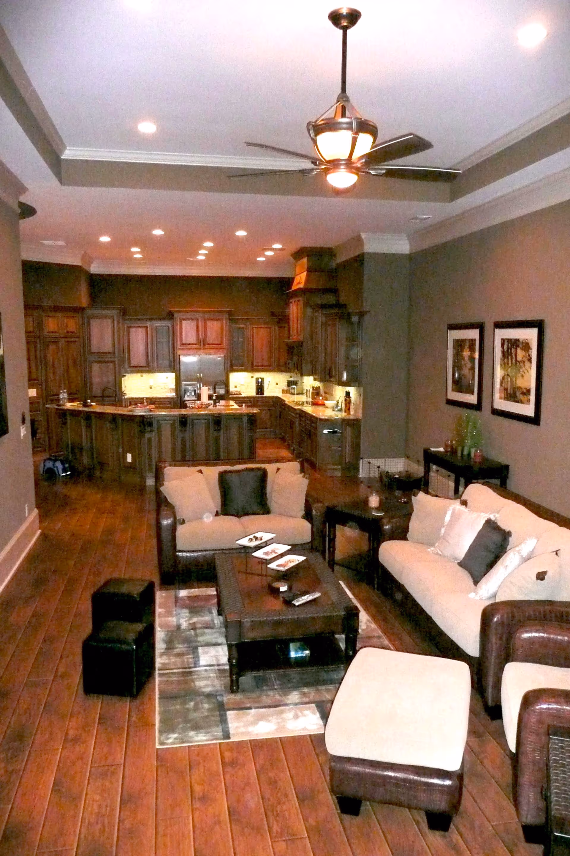 A cozy living room area with beige and brown cushioned sofas and ottomans arranged around a dark wooden coffee table on a patterned rug. The room has wooden flooring and neutral-colored walls with two framed pictures. In the background, there is a kitchen with dark wooden cabinets, a refrigerator, and under-cabinet lighting. A ceiling fan with lights is mounted on the ceiling.