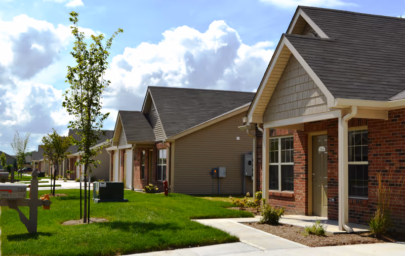 Row of single-story residential buildings with brick and siding exteriors, small front lawns, young trees, and a sidewalk under a partly cloudy sky.