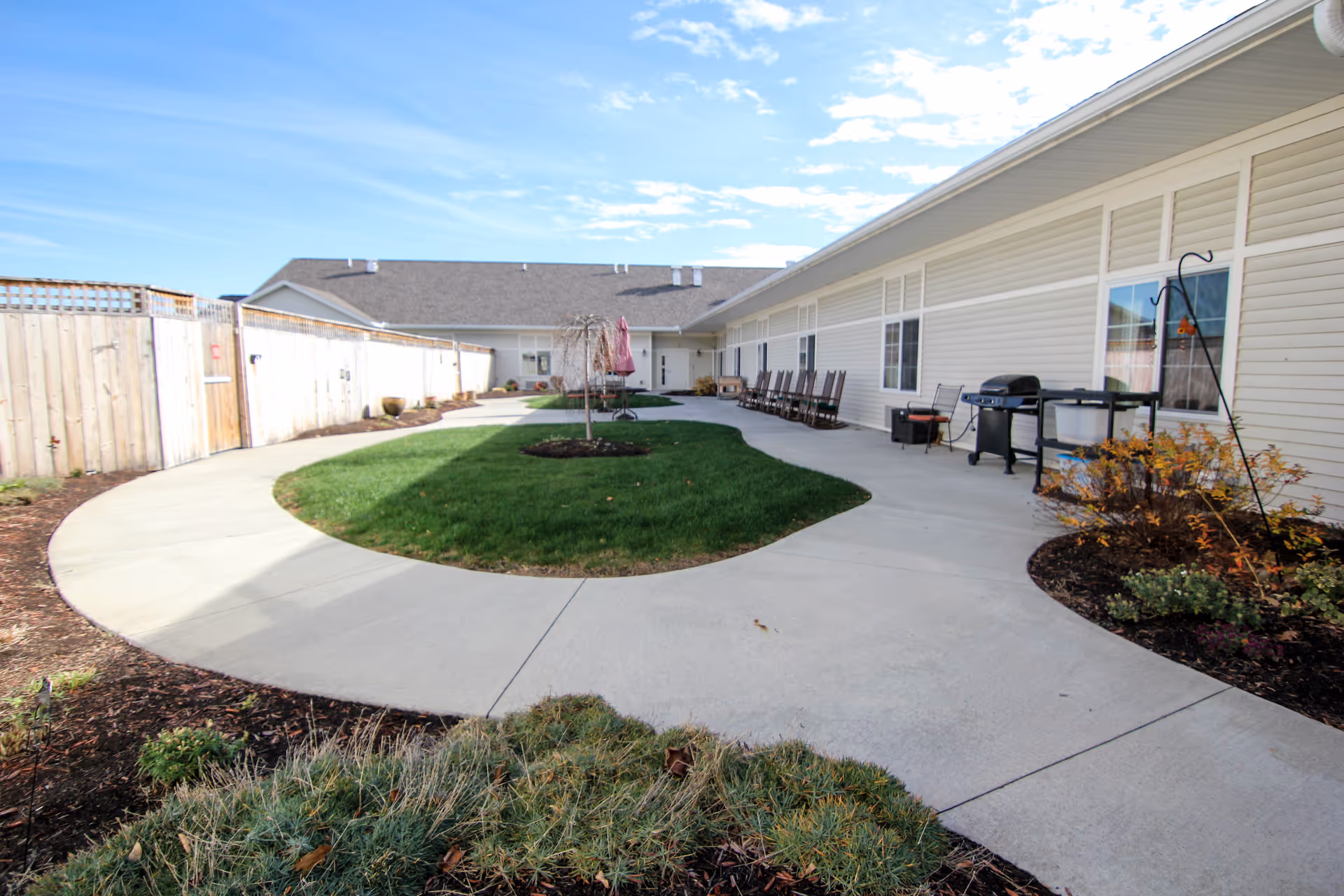 Outdoor courtyard area with a curved concrete walkway surrounding a grassy island with a small tree. The courtyard is bordered by a light-colored building with several windows and a row of rocking chairs. There is a barbecue grill and some patio furniture along the building. The sky is partly cloudy with blue patches.
