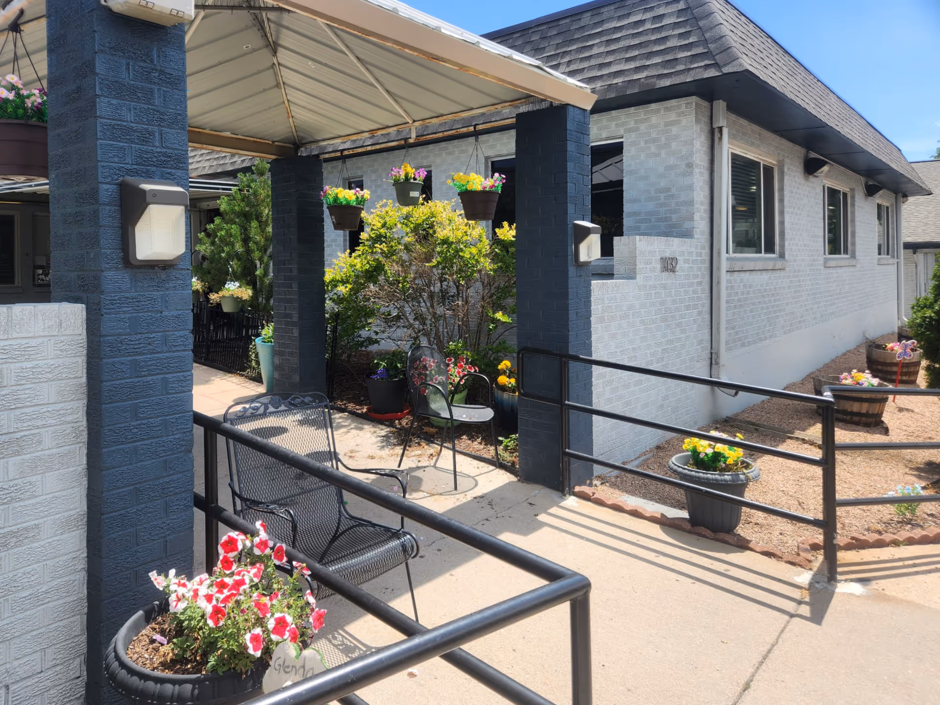 Outdoor entrance area of a building with a covered walkway supported by dark blue pillars. There are several flower pots with colorful flowers, hanging flower baskets, and a metal chair along the pathway. The building has light gray brick walls and multiple windows. A black metal handrail lines the walkway.