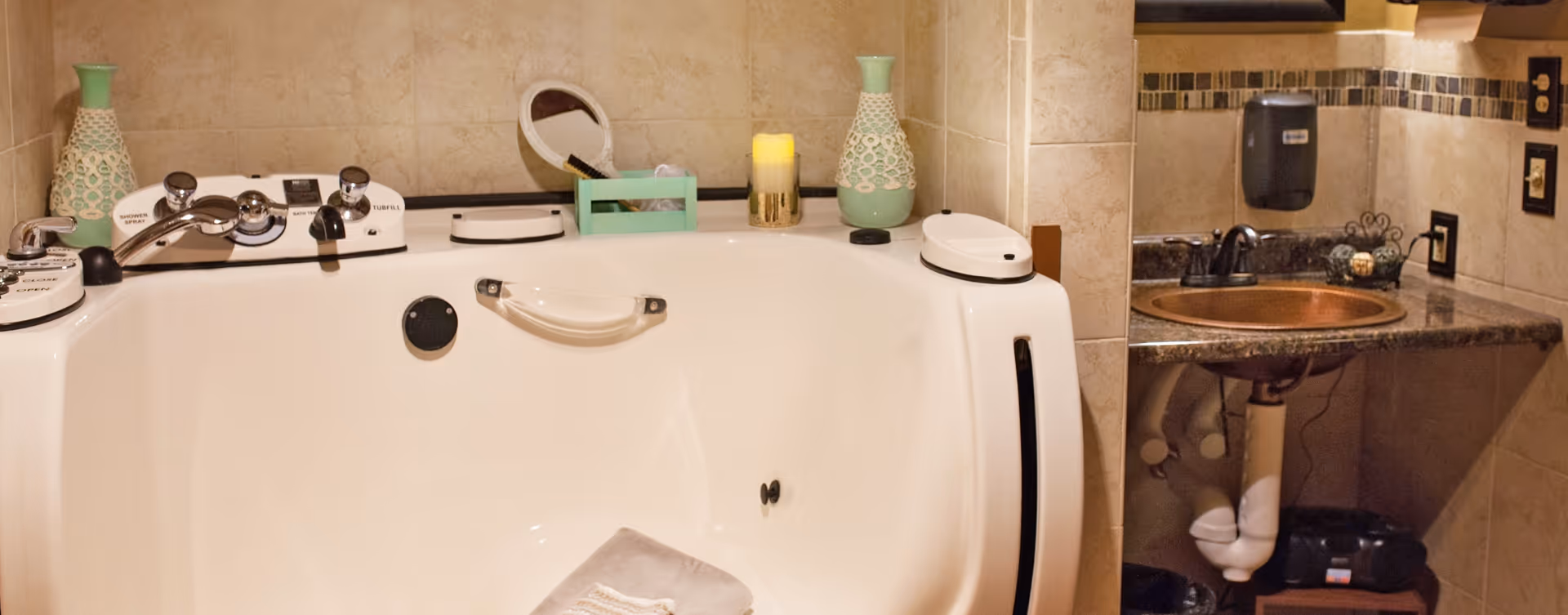 A bathroom featuring a large white bathtub with various controls and handles, a folded towel placed inside the tub, decorative vases and a candle on the tub ledge, and a sink with a dark countertop and a soap dispenser mounted on the wall.