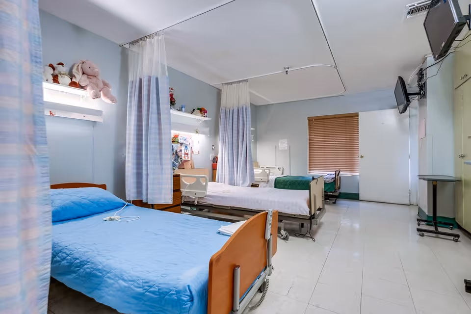 Shared nursing facility room with several hospital beds separated by privacy curtains and a wall-mounted TV.