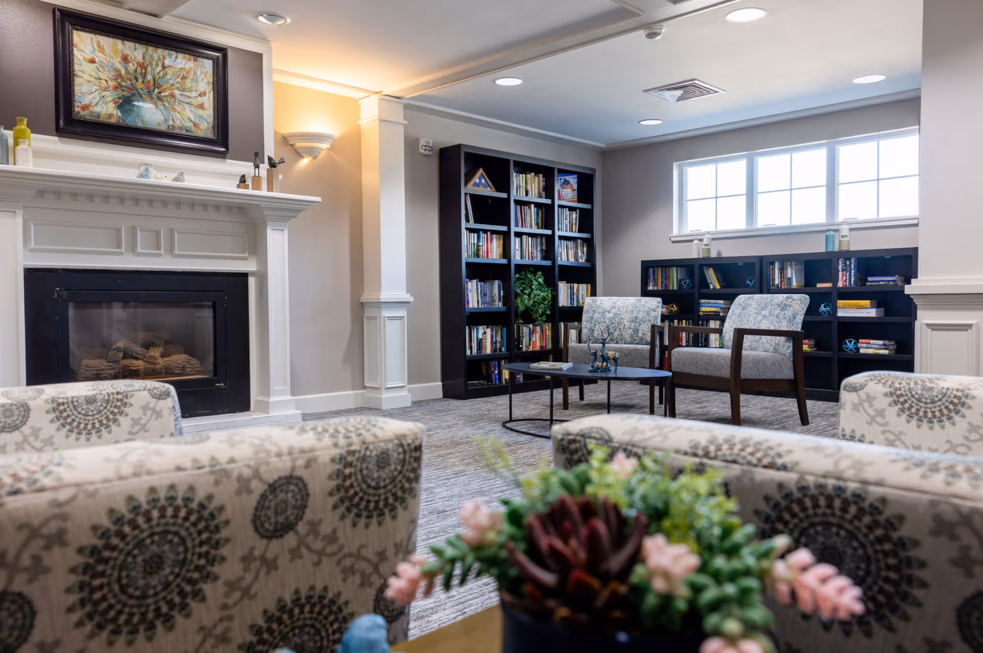 A cozy living room area with patterned armchairs, a fireplace with a painting above it, a coffee table with decorative items, and bookshelves filled with books against the walls. There is a window letting in natural light and a potted plant in the foreground.