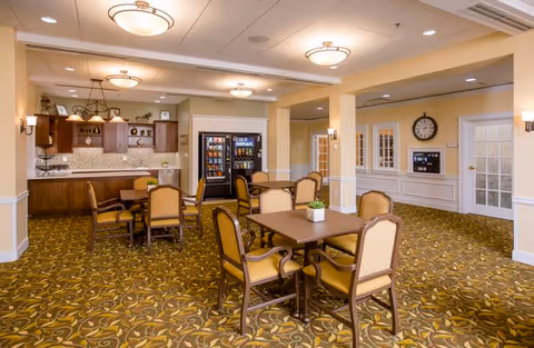 A spacious dining area in a senior living facility featuring several tables with cushioned chairs. The room has warm lighting with ceiling fixtures and wall sconces. In the background, there is a kitchenette with wooden cabinets, a countertop, and a vending machine. The carpet has a decorative pattern, and there is a clock on the wall near a set of glass-paneled doors.