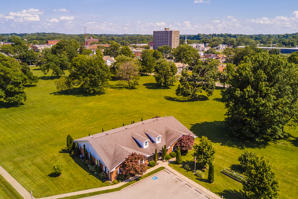Aerial view of a single-story brick building surrounded by a large green lawn with trees. The building has a brown shingled roof with dormer windows and is located near a parking lot with a handicapped parking space. In the background, there are more trees, residential houses, and a taller building under a blue sky with scattered clouds.