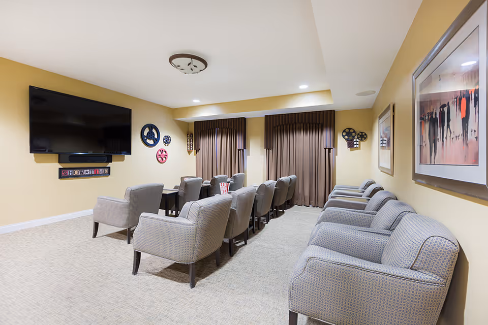 A cozy media room with multiple upholstered armchairs arranged in rows facing a large flat-screen TV mounted on a beige wall. The room has carpeted flooring, brown curtains covering the windows, and decorative film reels and framed artwork on the walls.