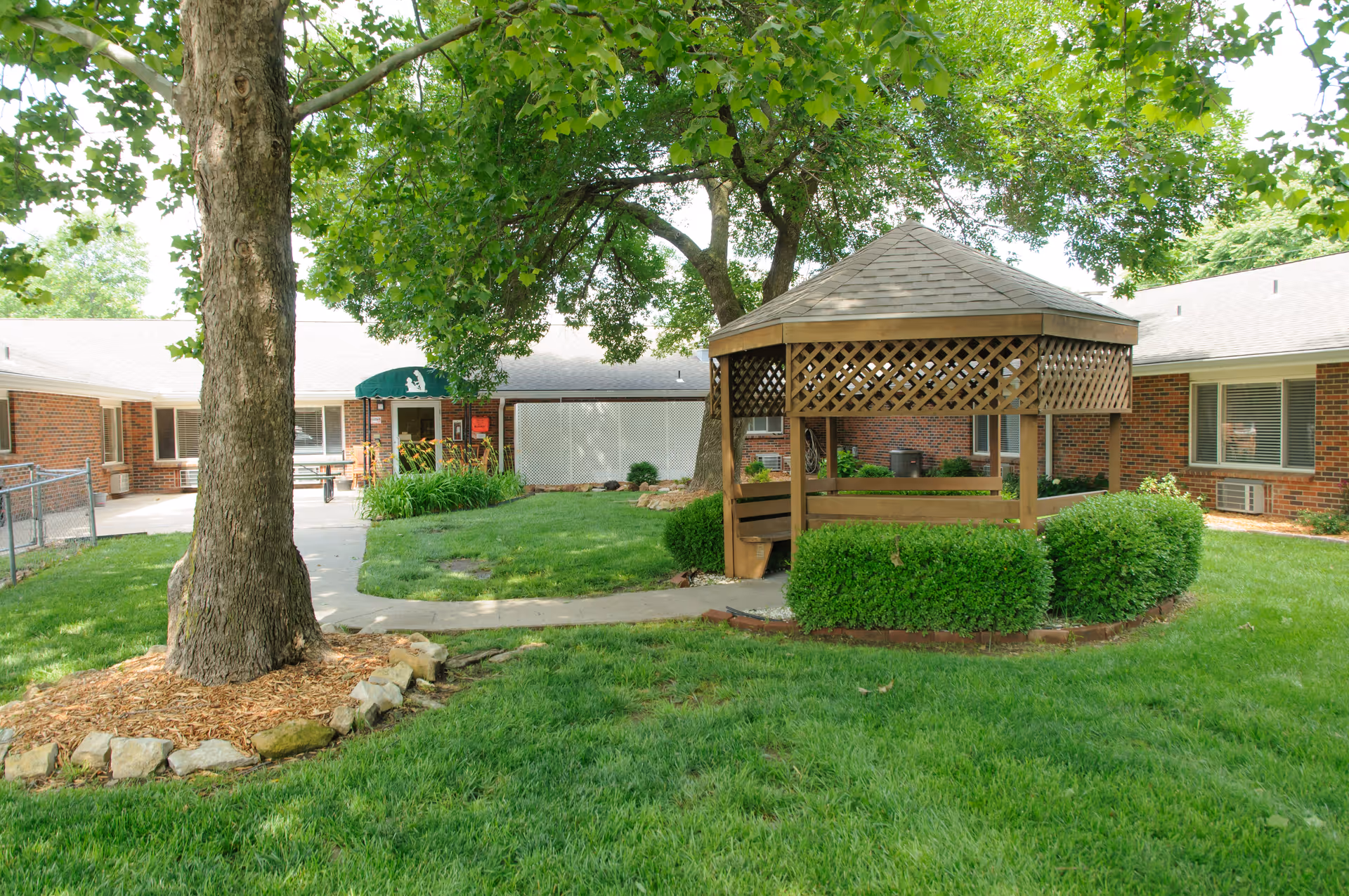 Outdoor courtyard area of a senior living facility with a large tree, a wooden gazebo surrounded by bushes, green grass, and a paved walkway. The brick building with windows and a green awning over the entrance is visible in the background.
