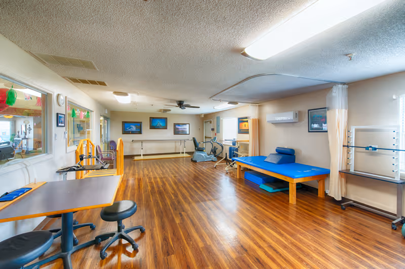 Spacious interior therapy and exercise room with wood floors, a blue treatment table, exercise bikes, tables and chairs, and wall-mounted artwork.