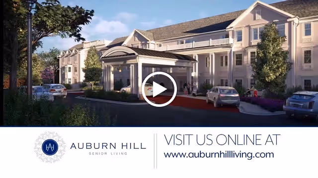 Exterior view of Auburn Hill Senior Living facility showing a large, multi-story building with a covered entrance driveway. Several cars are parked near the entrance, and there are trees and landscaping around the building under a partly cloudy sky.