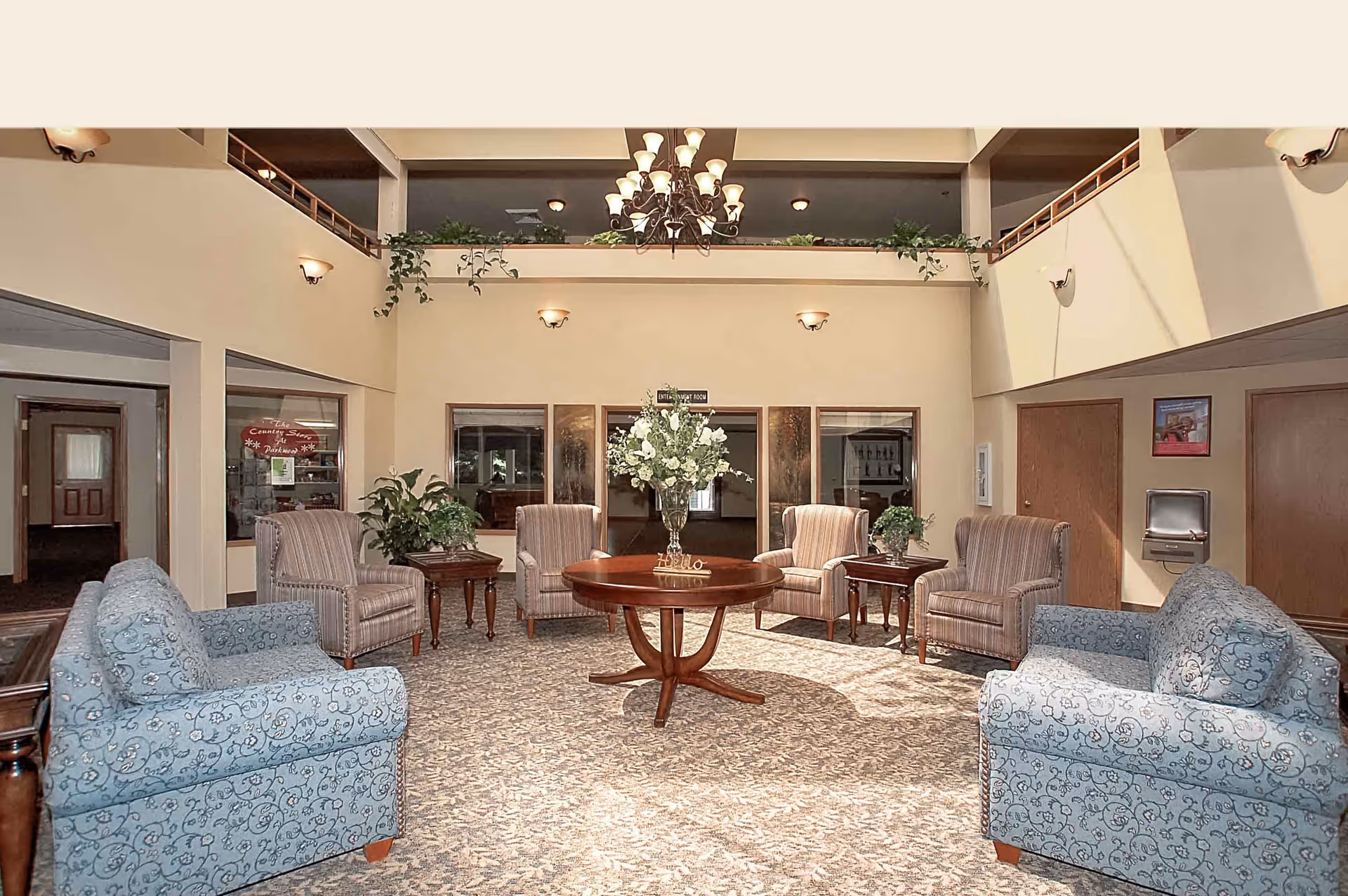 A spacious and well-lit common area in a senior living facility with high ceilings and a chandelier. The room features a round wooden table with a floral centerpiece in the middle, surrounded by four striped armchairs and two blue patterned sofas. There are side tables with plants, wall sconces providing lighting, and a balcony railing visible on the upper level. Doors and windows lead to other rooms in the background.