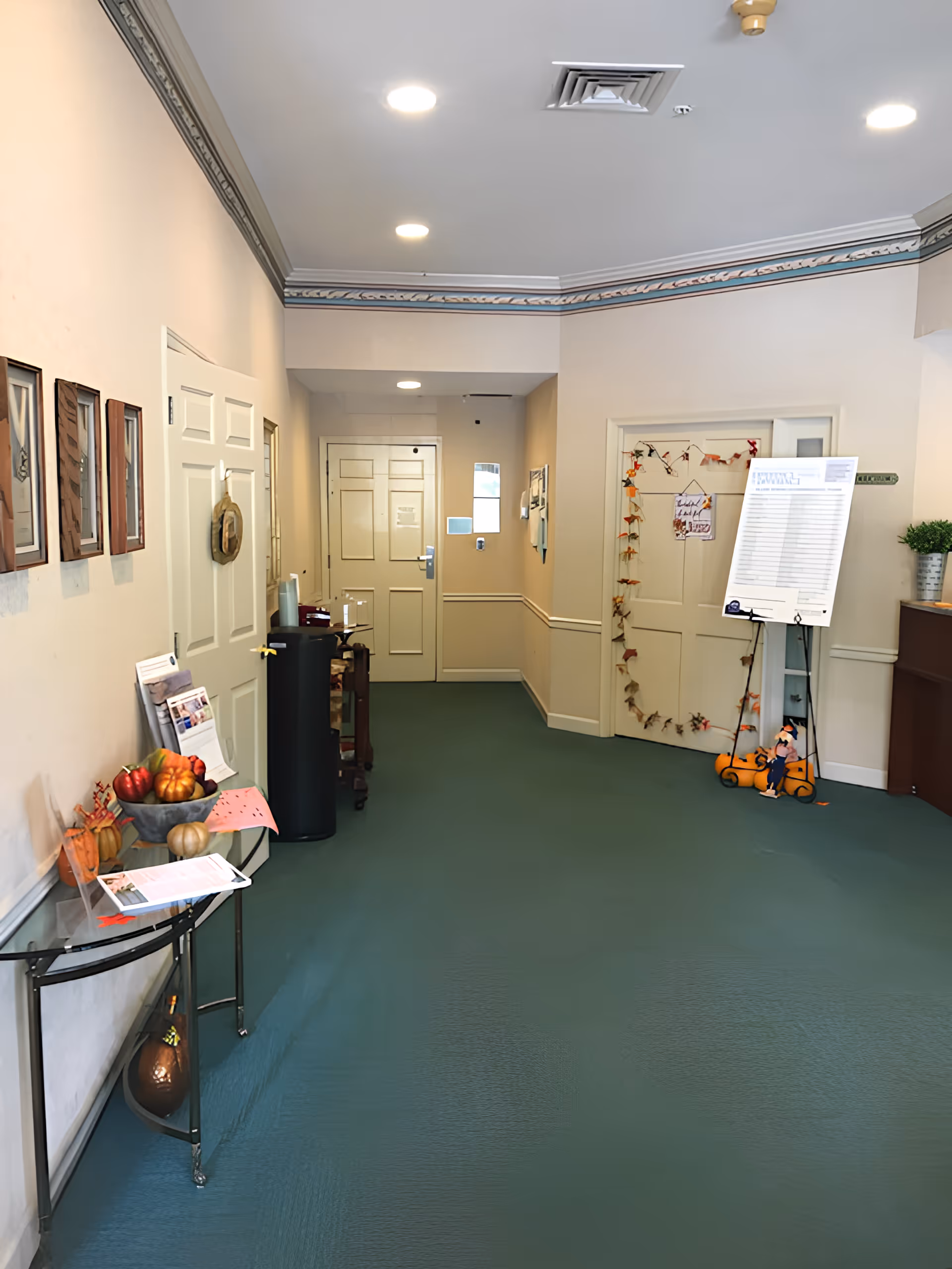 Interior hallway of Arden Courts - ProMedica Memory Care Community with teal carpet, beige walls, and white doors. The hallway features framed pictures on the left wall, a small table with decorative pumpkins and papers, a water dispenser, and a bulletin board on the right side near the end. The ceiling has recessed lighting and decorative molding.