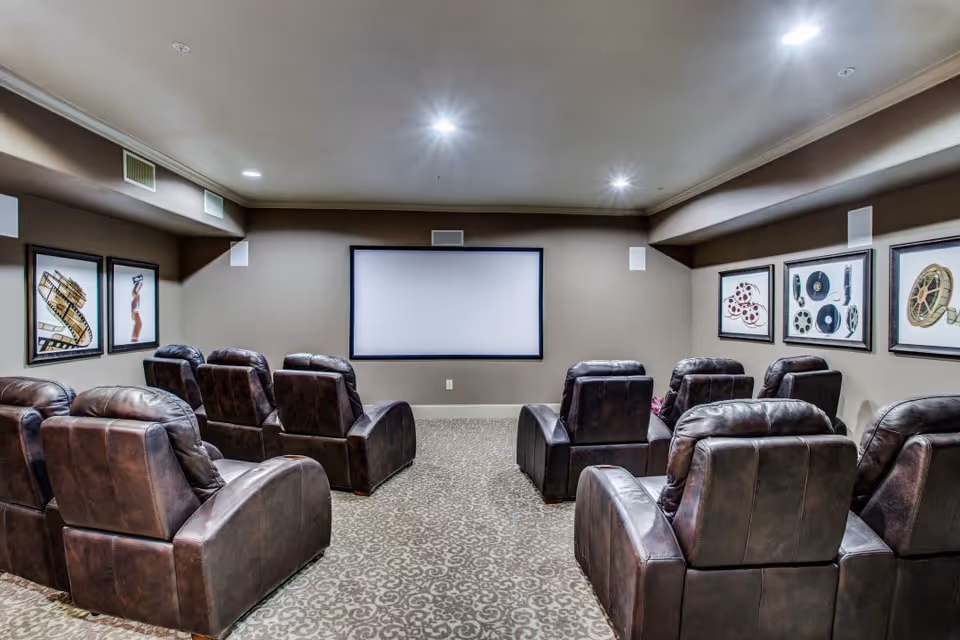 A small theater room with two rows of dark brown leather recliner chairs facing a blank projection screen on a beige wall. The room has patterned carpet flooring and framed artwork featuring film reels and movie-related images on the walls.