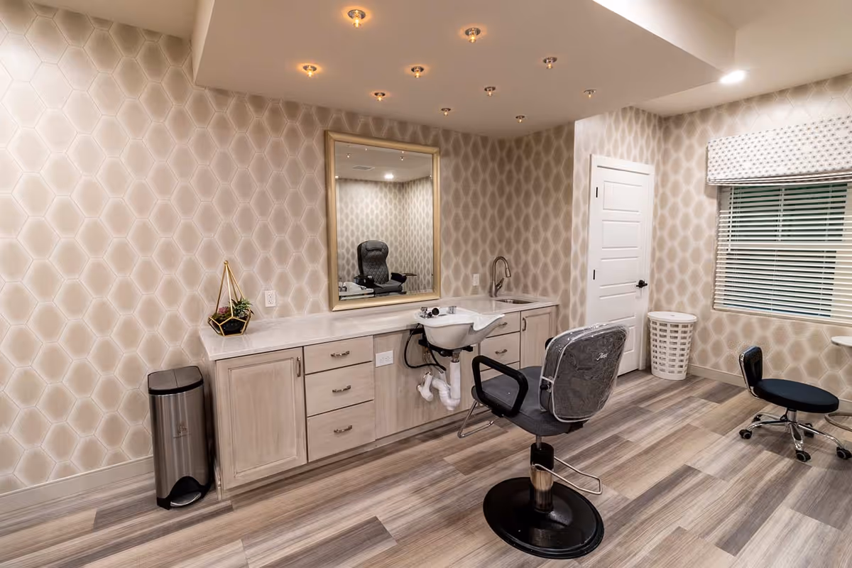 Interior of a senior living facility's hair salon area featuring a salon chair in front of a wash basin and a large mirror mounted on a patterned wall. The room has light wood flooring, a trash can, a small plant on the counter, a window with blinds, and a rolling stool.