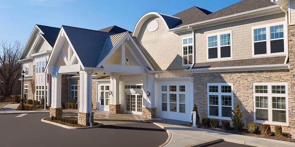 Front exterior view of The Bristal Assisted Living at Lake Success building with a covered entrance, stone and siding facade, multiple windows, and a driveway.