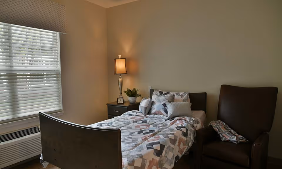 A cozy bedroom with a single bed covered in a patterned quilt and multiple pillows. Next to the bed is a nightstand with a lamp, a small clock, and a potted plant. A large window with blinds allows natural light to enter the room. There is also a dark brown armchair with a folded blanket draped over one arm.