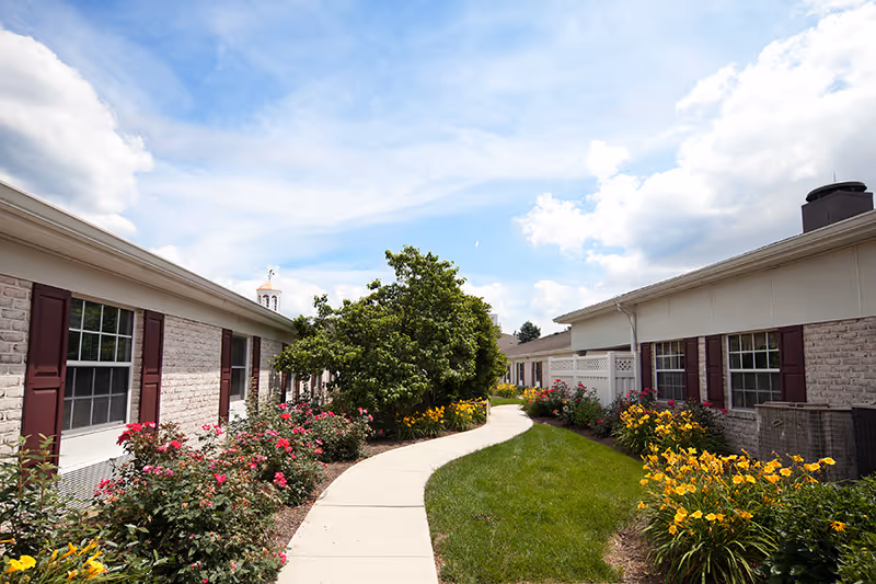 Curved concrete pathway between two single-story brick buildings with maroon shutters, surrounded by green grass, blooming yellow and pink flowers, and a large leafy tree under a partly cloudy blue sky.
