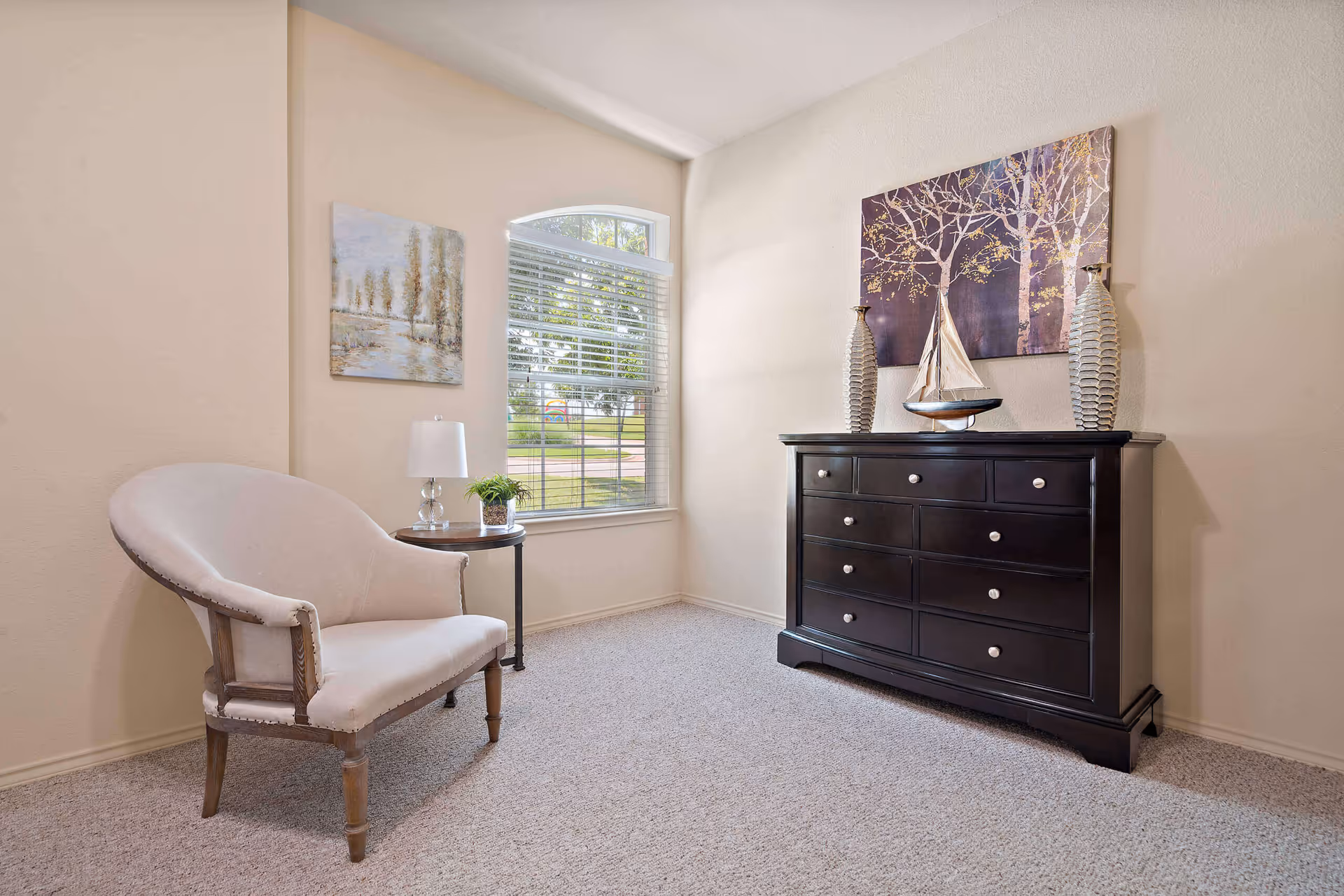 A cozy corner of a room with a beige upholstered armchair, a small round wooden side table with a lamp and a small plant, a large window with white blinds letting in natural light, and a dark wooden dresser decorated with two tall vases and a model sailboat. Two paintings hang on the walls, one depicting trees by a river and the other showing trees with golden leaves.