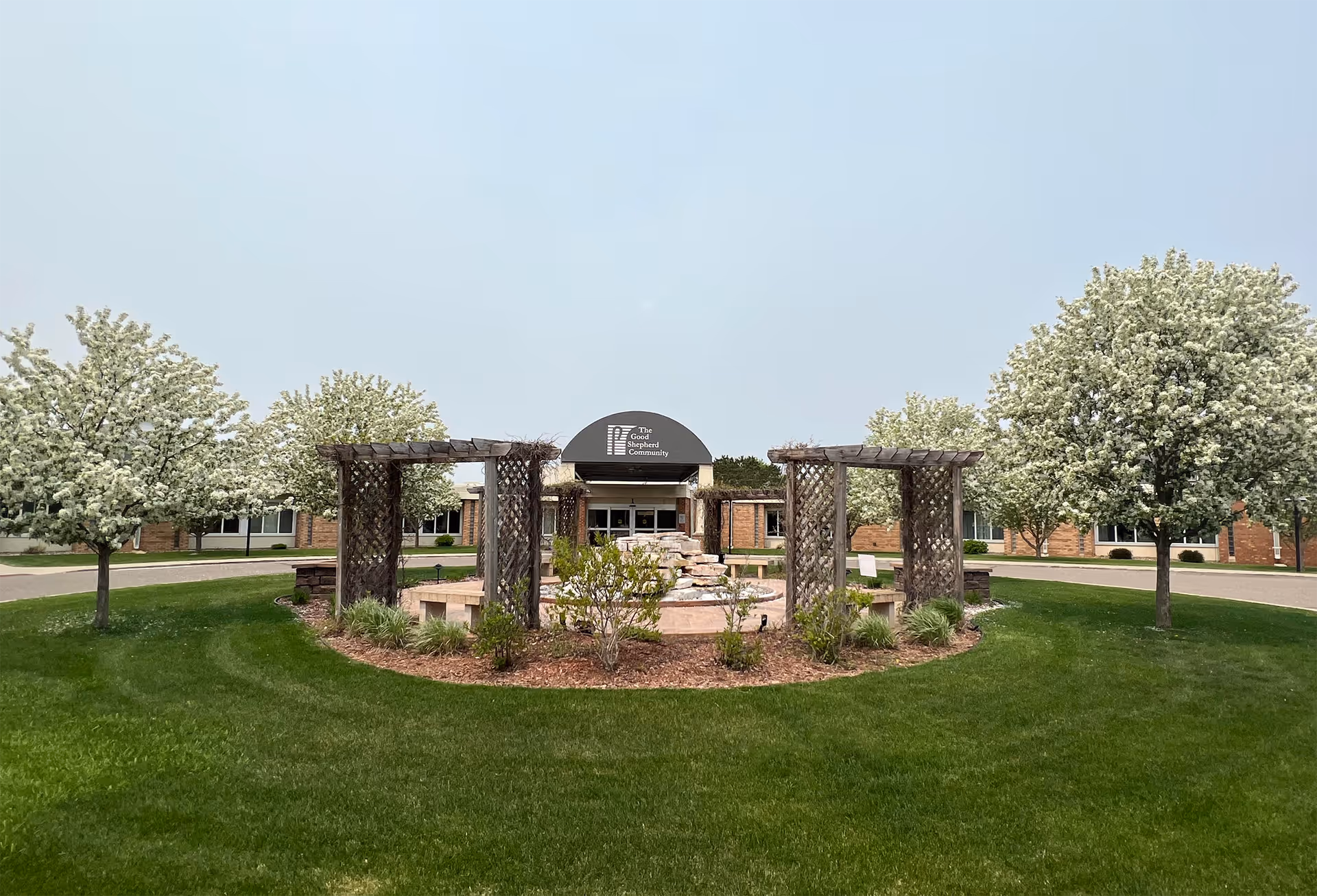 Front entrance of Good Shepherd Community with a landscaped circular courtyard, wooden pergolas, blooming trees, and the facility sign above the door.