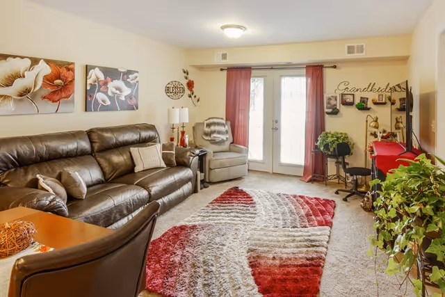 A cozy living room with a dark brown leather sofa, a gray armchair, and a colorful red, white, and gray patterned rug on a carpeted floor. The room has beige walls decorated with floral artwork and wall hangings, including one that says 'Grandkids'. There are red curtains on the glass doors leading outside, and several green plants add a touch of nature to the space.