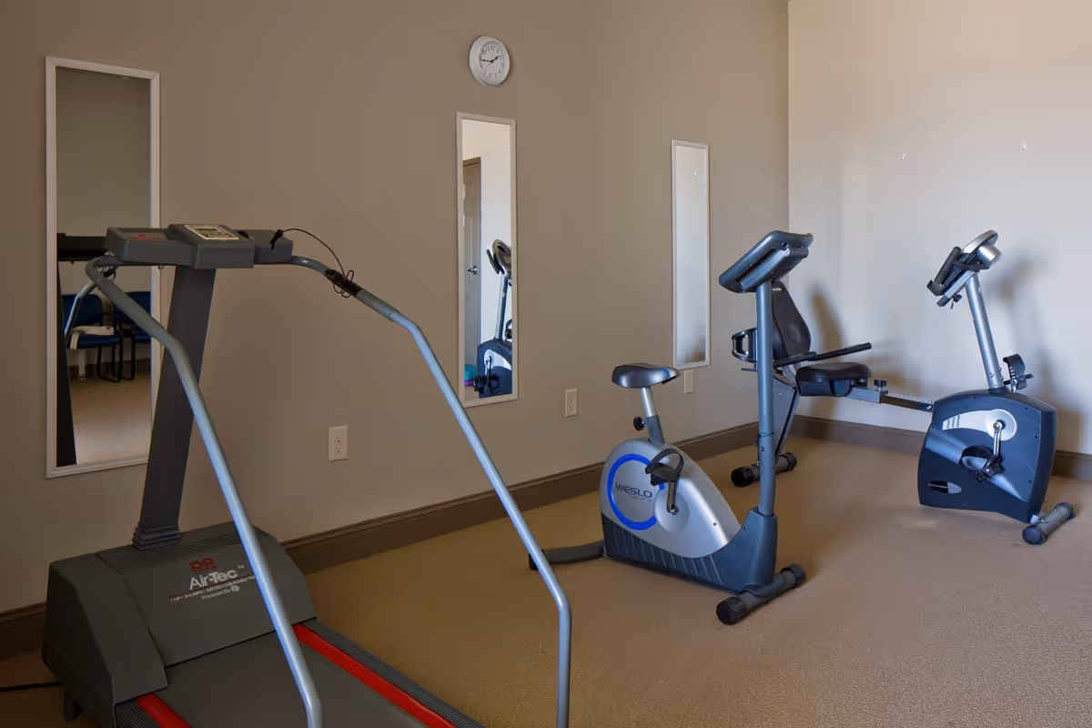 A small exercise room with a treadmill and two stationary bikes against beige walls with two vertical mirrors and a clock above one mirror.