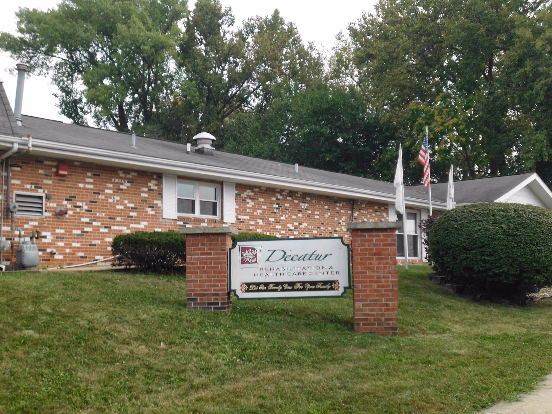 Exterior view of Decatur Rehabilitation Health Care Center showing a brick building with a sign in front on a grassy area. The sign reads 'Decatur Rehabilitation & Health Care Center' with the tagline 'Let Our Family Care For Your Family'. There are bushes, trees, and an American flag near the building.
