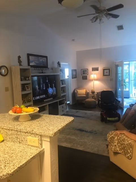 Interior view of a senior living facility common area with a granite countertop in the foreground holding a fruit bowl. The room includes a TV on a wooden entertainment center, a ceiling fan, a lamp on a side table, armchairs, a mobility scooter, and framed pictures on the walls. There is natural light coming through glass doors leading outside.