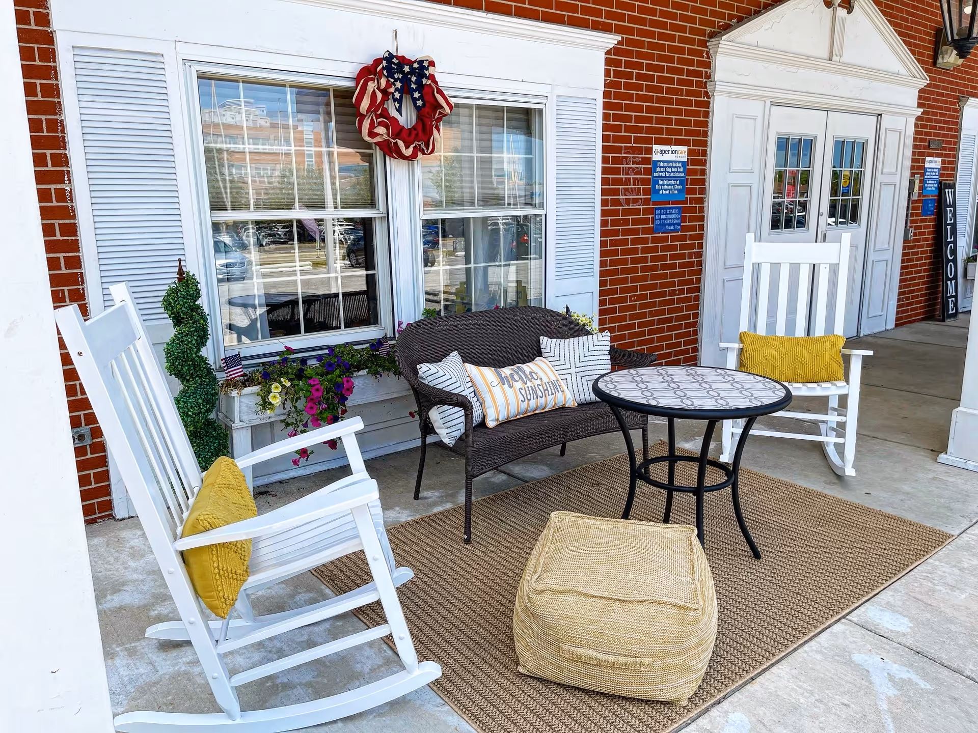 Outdoor seating area in front of a brick building with white trim. The area includes two white rocking chairs with yellow cushions, a dark wicker loveseat with multiple pillows including one that says 'Hello Sunshine', a round table with a patterned top, and a beige pouf on a woven rug. A patriotic wreath hangs on the window behind the seating, and there are flower boxes with colorful flowers beneath the window. The entrance door is white with glass panels and a 'WELCOME' sign is visible nearby.
