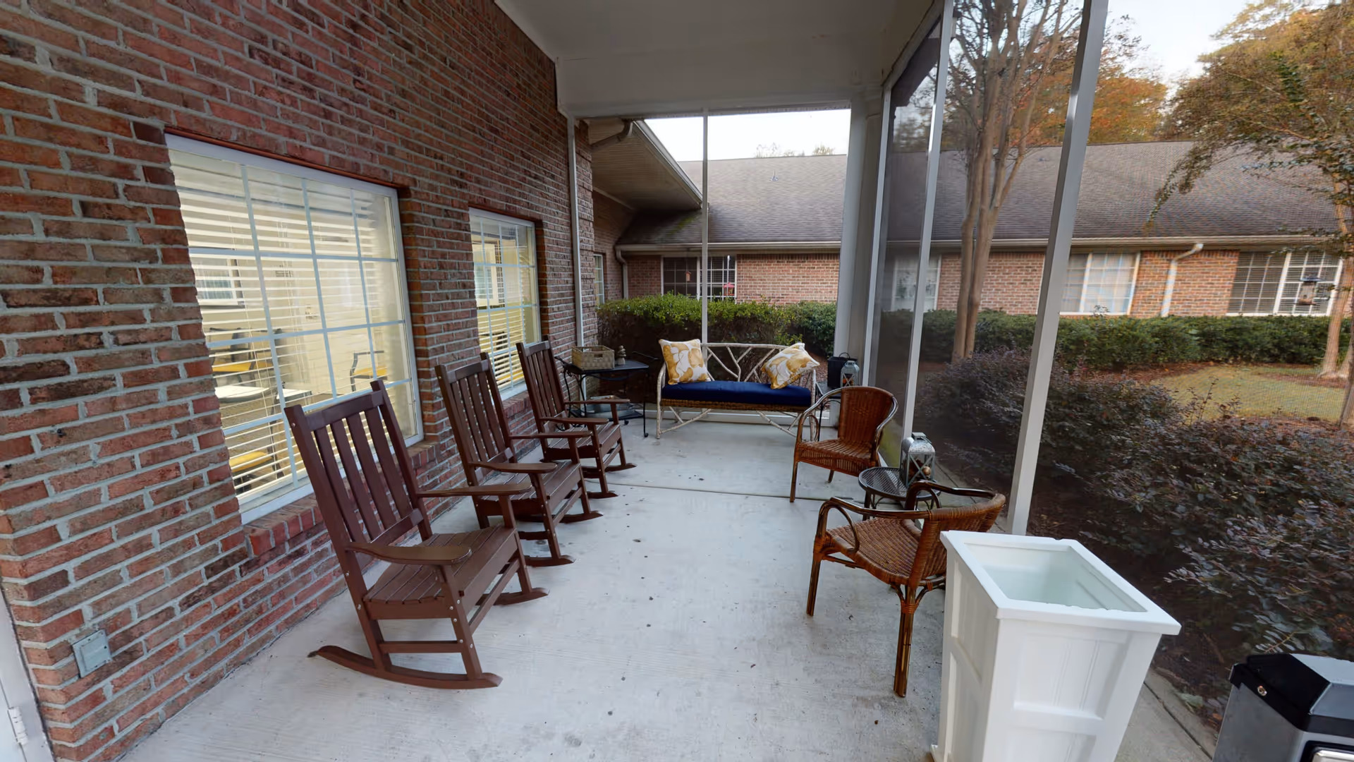 A screened outdoor patio with wooden rocking chairs, wicker seats, and a cushioned bench along a brick wall.