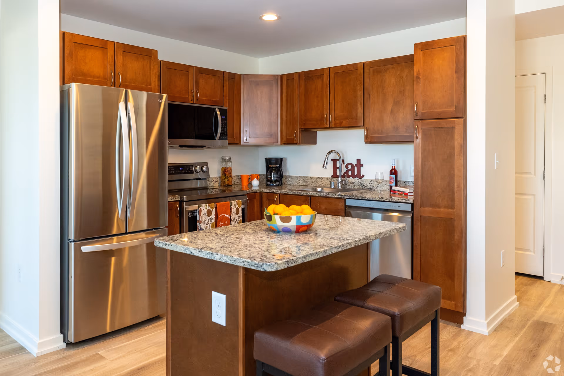 Modern kitchen with wooden cabinets, stainless steel refrigerator, microwave, stove, and dishwasher. Granite countertop island with a bowl of oranges and two brown cushioned stools. The word 'Eat' is displayed on the countertop near the sink.