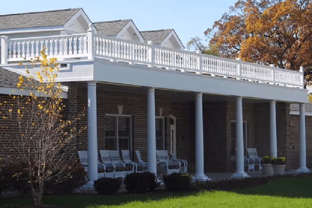 Exterior view of a brick building with a white porch supported by multiple white columns. Several white rocking chairs are arranged on the porch. There is a small tree with yellow leaves and green grass in the foreground, and autumn-colored trees in the background.