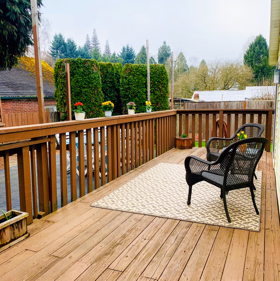 A wooden outdoor deck with two black wicker chairs placed on a patterned beige rug. The deck is surrounded by a wooden railing with several potted flowers on top. In the background, there are tall green bushes, trees, and a neighboring building with a moss-covered roof.
