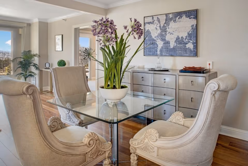A bright dining area with a glass table surrounded by four ornate beige upholstered chairs. A white pot with tall purple flowers is centered on the table. Behind the table is a light-colored wooden sideboard with drawers, topped with decorative items including a glass decanter and a stack of red plates. A framed world map hangs on the wall above the sideboard. The room has wooden flooring and large windows letting in natural light, with a view of an outdoor balcony and cityscape in the background.